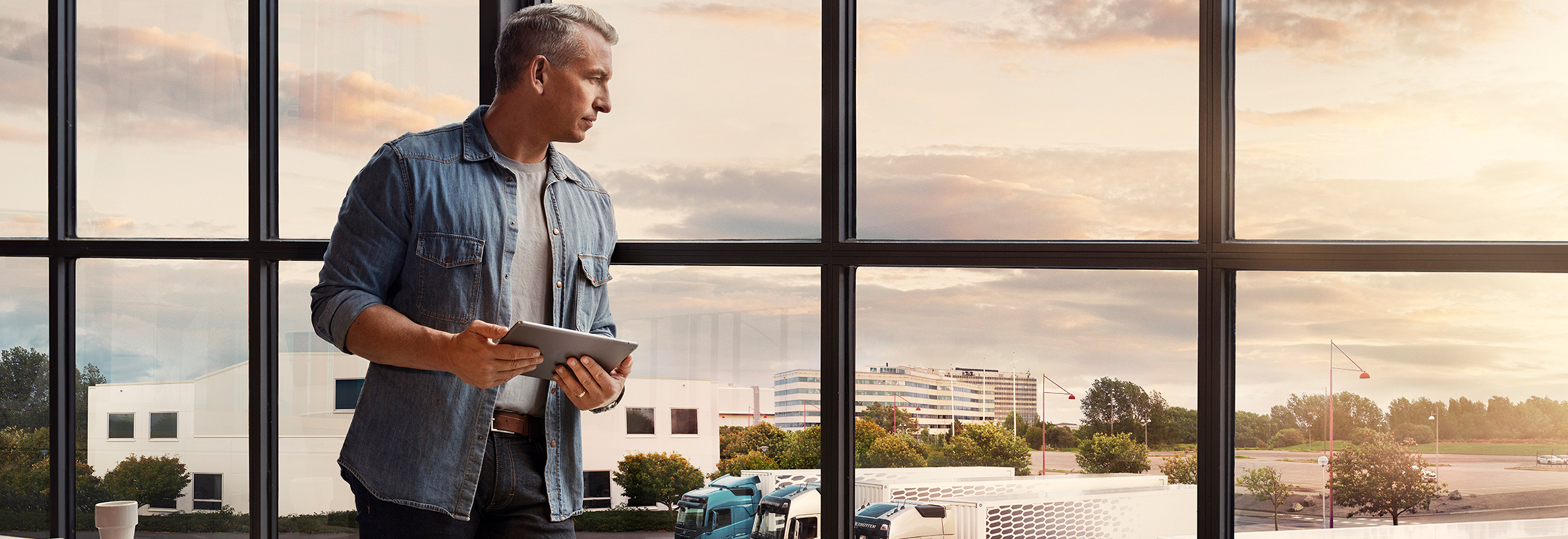 Muž držící tablet stojí u okna a dívá se dolů na svůj vozový park