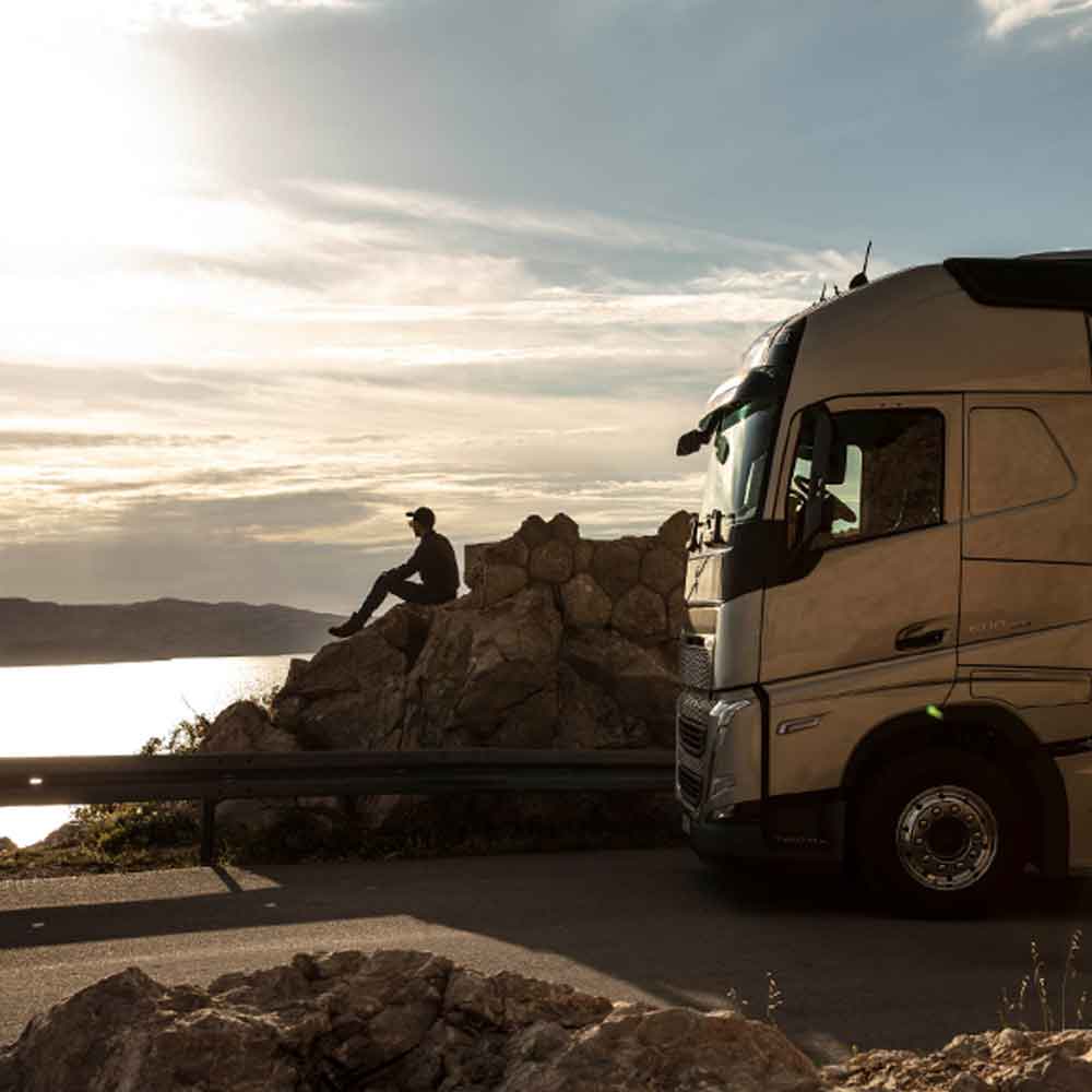 Řidič sedí na skále a při západu slunce se dívá na moře, vedle něj stojí Volvo FH